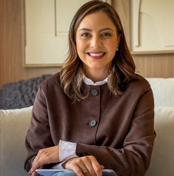 Dani Padilha sorrindo, de blazer marrom, sentada no sofá